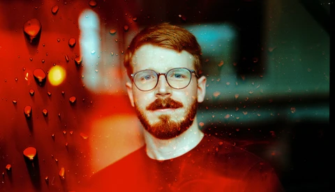 Man wearing glasses and red shirt photographed through rain-covered glass with warm and cool color cast.