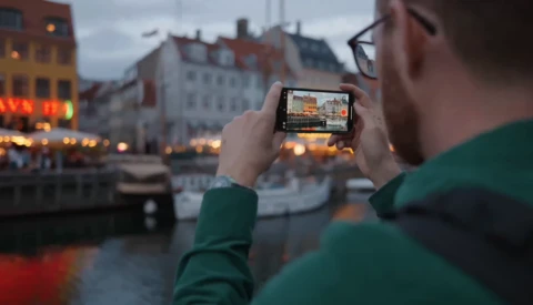 Photographer in green jacket holding smartphone horizontally while framing a canal-side European cityscape during golden hour.