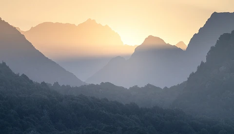 Sun rising between forested mountain peaks shrouded in layered mist and haze.