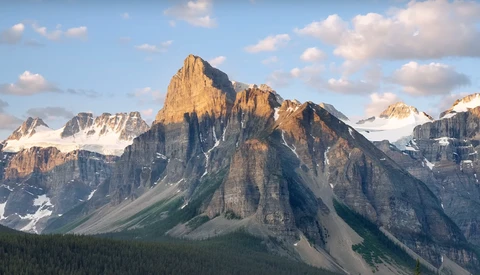 Dramatic mountain peak with vertical columnar rock formations, snow patches, and forested valley below under partly cloudy sky.