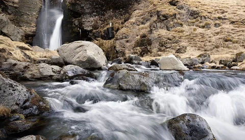 Mountain stream with cascading white water flowing around moss-covered rocks and boulders, with a waterfall visible in the background.