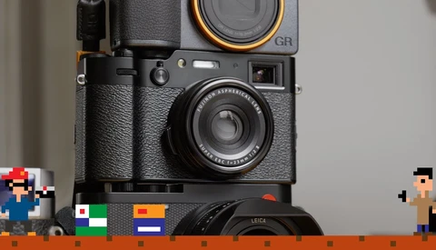 Stack of vintage cameras including a Ricoh GR film camera and Leica lens with pixel art figures and colored film boxes in foreground.