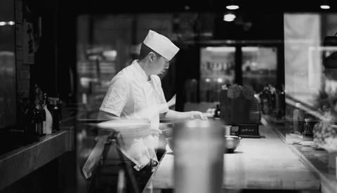 Chef in white uniform and head covering working at counter in commercial kitchen.