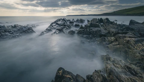 Rocky coastal outcrops shrouded in silky mist with hills visible across calm water.