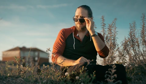 Man in orange shirt and sunglasses crouching in a field during golden hour light.
