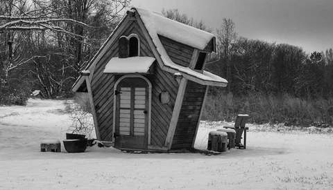 Tilted wooden structure resembling a house with snow-covered roof and surrounding winter landscape.