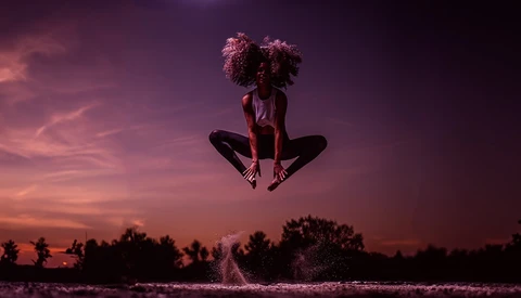 Person with curly hair jumping above water during purple sunset, captured mid-air in silhouette.