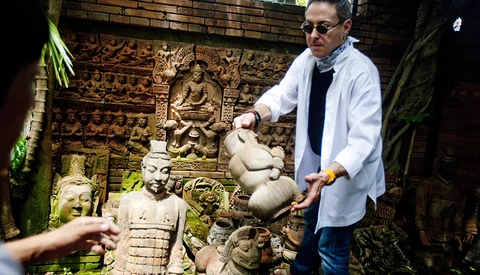 Man in light blue shirt gesturing toward stone sculptures in an overgrown courtyard with carved relief panels.