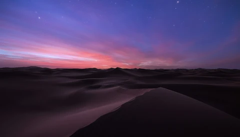 Desert dunes silhouetted against a vibrant twilight sky with purple and pink hues and visible stars.