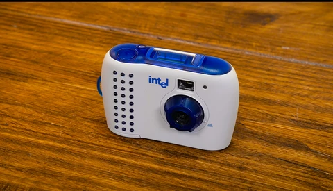 Intel-branded compact point-and-shoot camera with white body, blue accents, and perforated speaker grille on wooden surface.