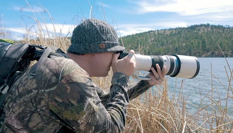 Wildlife photographer with telephoto lens capturing images by a forested lake shore.