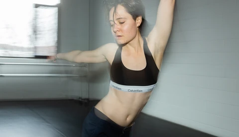 Woman in sports bra performing a side stretch against a gray wall with natural window light.
