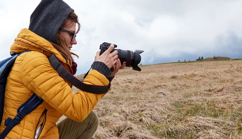 Photographer in yellow jacket and hood holding DSLR camera while crouching in open field.