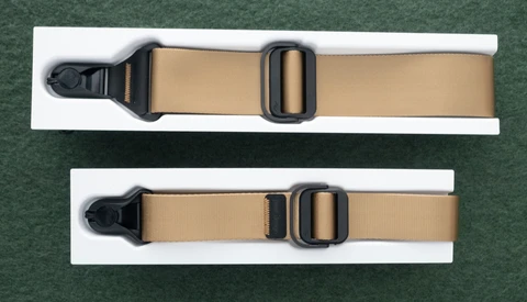Two tan webbing straps with black plastic side-release buckles displayed on white cards against a green background.