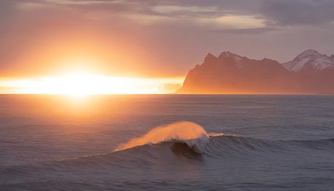 Ocean wave with golden spray backlit by sunset, snow-capped mountain silhouetted on horizon.