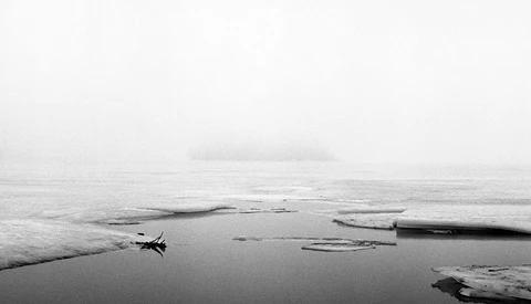 Minimalist black and white landscape with a lone boat on still water surrounded by driftwood logs in mist.