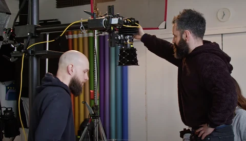 Two men adjusting a professional camera mounted on a rig in a studio, with colored gels visible in the background.