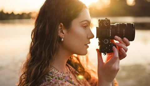 Woman holding a mirrorless camera to her face during golden hour, silhouetted against a body of water.