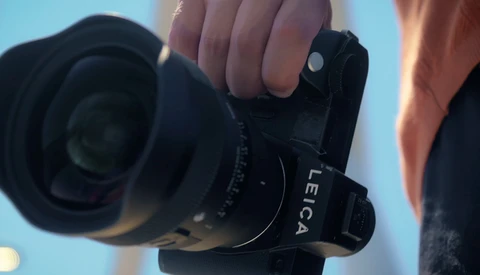 Photographer holding a mirrorless camera with a wide-angle lens against a blue sky background.