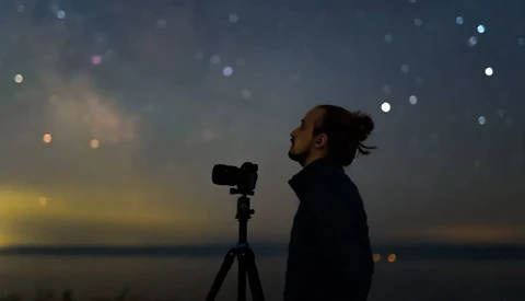 Photographer silhouetted against twilight sky, aiming camera on tripod toward starry horizon.