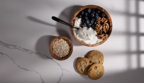 Overhead flat lay of breakfast ingredients in wooden bowls: yogurt with blueberries and granola, oats, and cookies on marble surface.