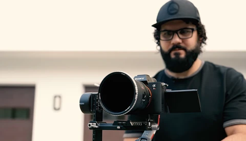 Photographer with a large telephoto lens mounted on a tripod head, wearing a baseball cap and glasses.