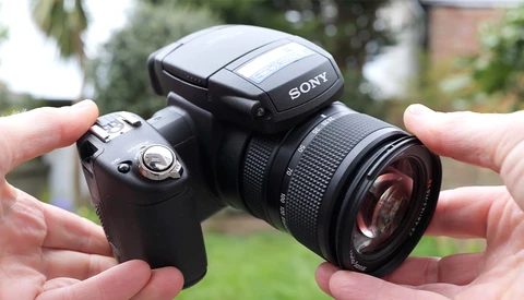 Sony camera with flash and telephoto lens held in both hands against outdoor background.