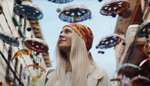 Woman with long platinum blonde hair and gold headwrap gazing upward surrounded by hanging decorative UFO sculptures.