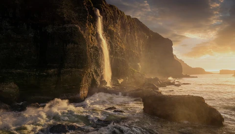 Waterfall cascading down a steep coastal cliff face during golden hour, with waves crashing on rocks below.