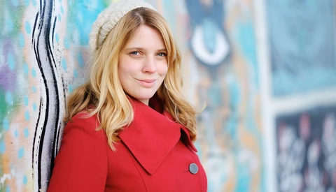 Portrait of a young woman with blonde hair wearing a red coat against a colorful textured wall.