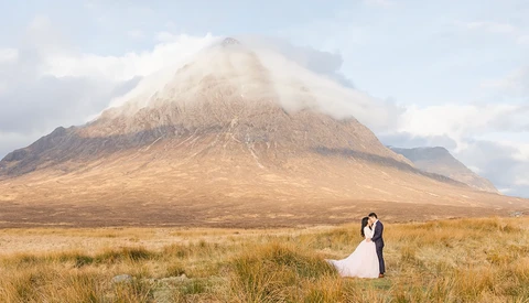 Bride and groom embracing in a vast golden grassland with a snow-capped volcanic mountain in the background.