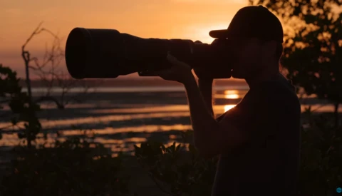 Silhouetted photographer with telephoto lens aimed at sunset over water during golden hour.