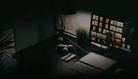 Photographer's desk setup with camera, laptop displaying image library, and monitor in moody side lighting.