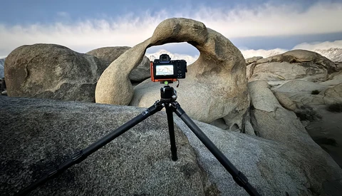 Smartphone mounted on tripod framing a natural rock arch against cloudy sky.