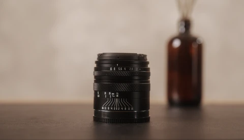 Black vintage lens with depth of field markings sits on wooden surface with brown glass bottle blurred in background.