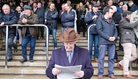Man in purple blazer and brown hat reading newspaper while standing on stadium steps surrounded by spectators.