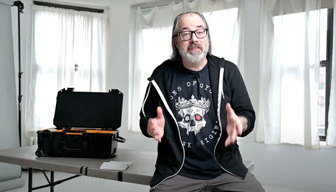 Man in glasses and black hoodie gestures while speaking in bright studio with equipment case on table.