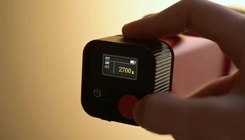Compact action camera with small LCD screen displaying 2700k color temperature reading, held in hand against warm-toned background.