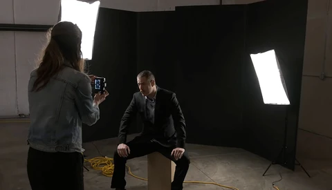 Photographer operating camera while interviewing man in suit in professional studio setup with large soft box lighting.