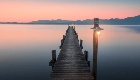 Wooden pier extending into still water at sunrise, with illuminated beacon post on right and mountain silhouettes beyond.