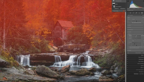 Waterfall flowing over rocks with autumn foliage and a red mill house in the background.