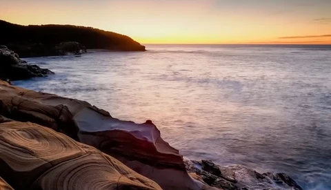 Rocky coastline at sunset with layered sandstone formations and calm ocean waters.