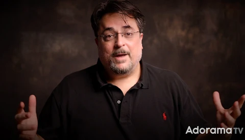 Man in black polo shirt gesturing while speaking against a dark textured background.