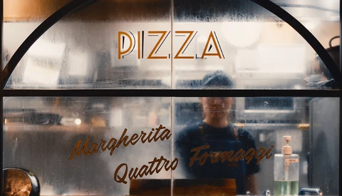 Pizza restaurant storefront window with neon signage and figure visible inside.