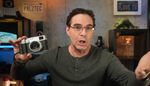 Man holding a vintage rangefinder camera while speaking to camera in a studio setting.