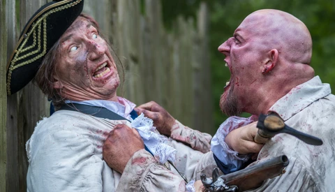 Two men in period costume engaged in a physical altercation near a wooden structure.