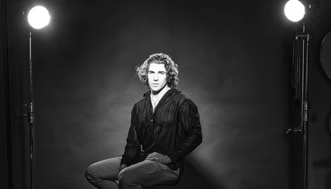 Man with curly hair sitting in studio lighting, wearing dark pinstriped shirt against dark background.