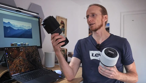 Man holding a cylindrical black device while gesturing toward a laptop displaying landscape photography.