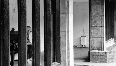 Person seated behind vertical wooden columns creating layered geometric composition in black and white.