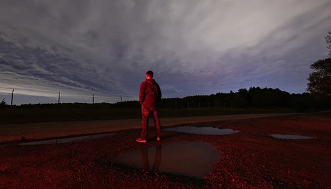 Figure in red clothing standing alone on wet red earth with water puddles under dramatic twilight sky.